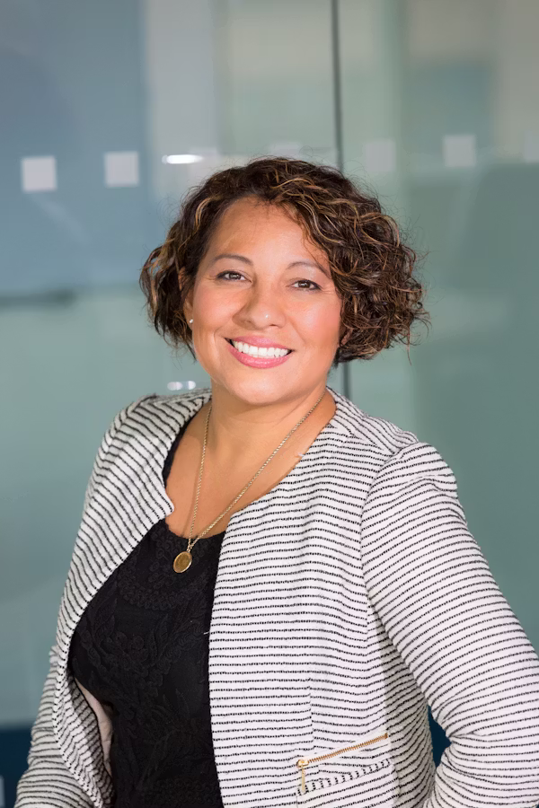 Curly haired woman smiling wearing black and white stripe blazer and black tops with gold. Necklace